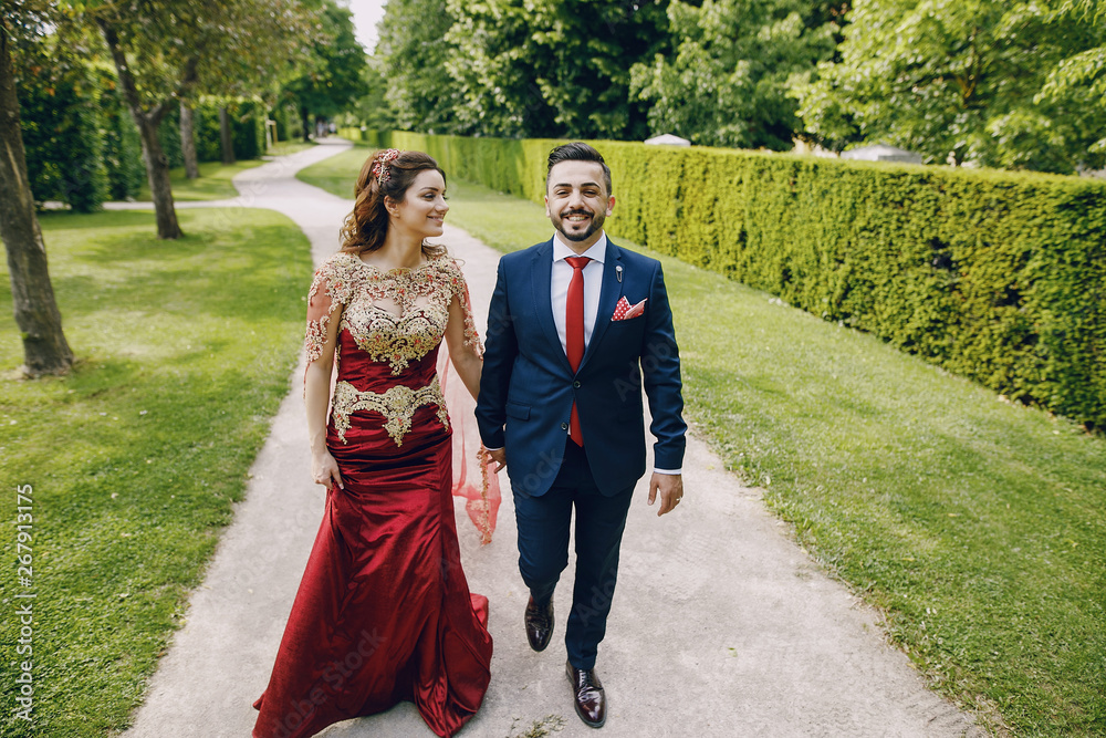 Fototapeta premium Beautiful woman in a long red dress walks around the city with her husband in a blue suit and with a beard