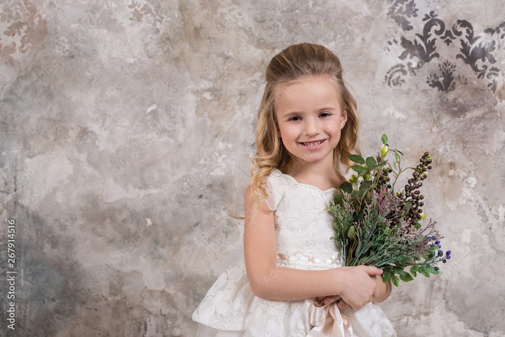 Portrait of a little attractive girl in a white dress with a bouquet in her hands against the background of a grunge wall.