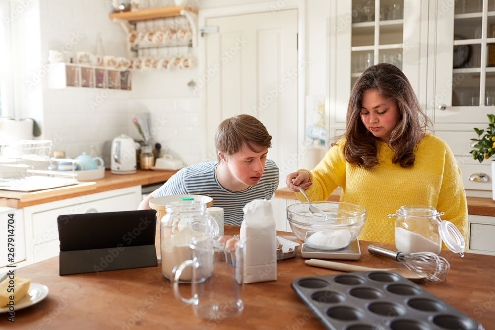 Young Downs Syndrome Couple Following Recipe On Digital Tablet To Bake Cake In Kitchen At Home