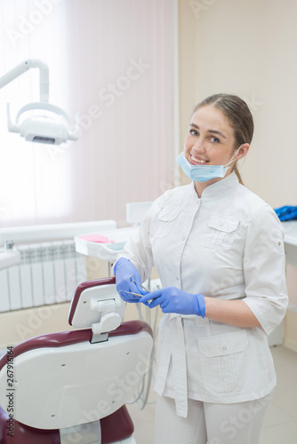 Attractive smiling female doctor in a white uniform at the workplace. Young beautiful female dentist in mask and gloves is standing in the office near the armchair with tools in her hands