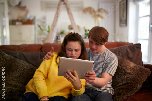 Young Downs Syndrome Couple Sitting On Sofa Using Digital Tablet At Home