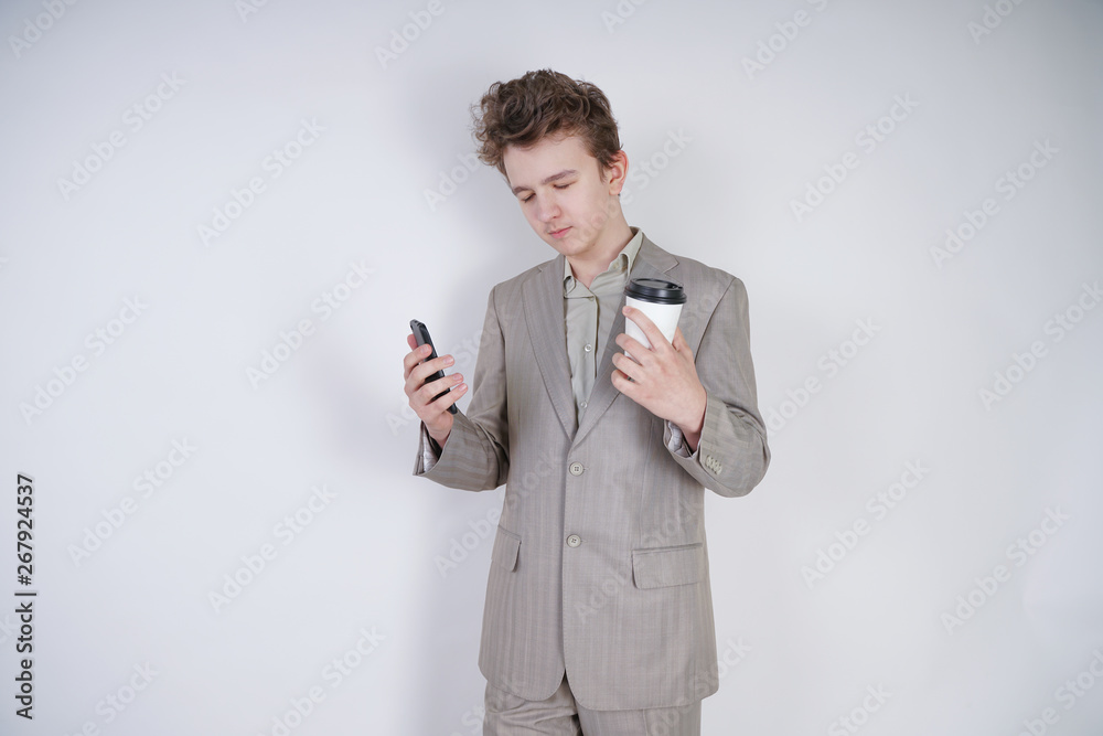 student teenager boy standing with closed eyes with coffee and smartphone. very tired young male wearing business suit on white studio background. student teenager boy standing with closed eyes with coffee and smartphone. very tired young male wearing business suit on white studio background.