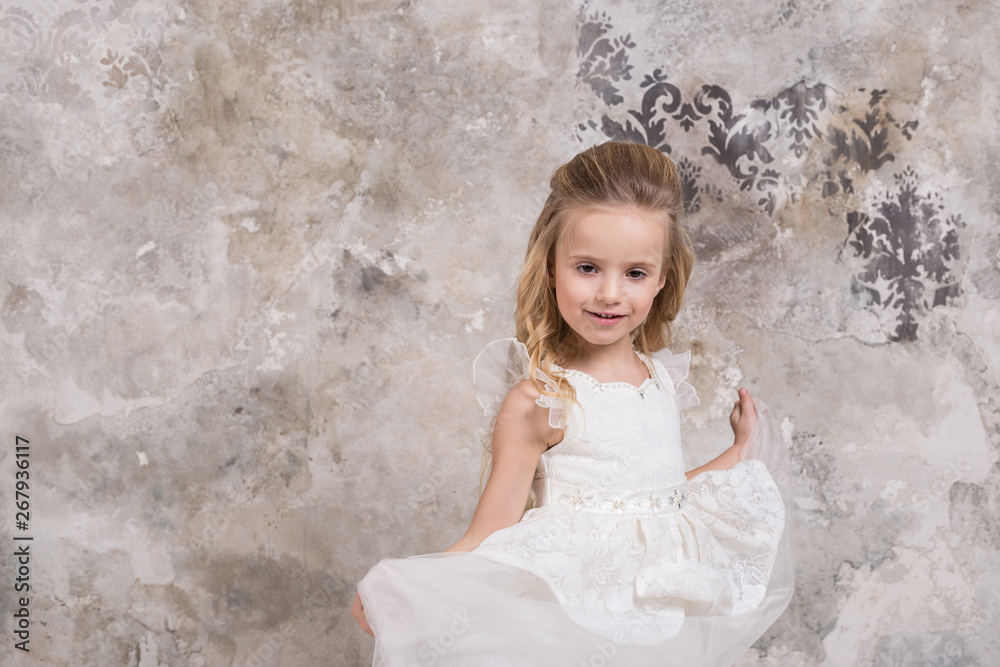 Portrait of a little attractive smiling girl in a white dress with curled hair against the background of a grunge wall. Portrait of a little attractive smiling girl in a white dress with curled hair against the background of a grunge wall.
