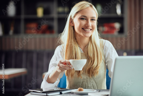 Charming smiling blonde businesswoman dressed smart casual sitting in cafeteria  drinking coffee and using laptop.