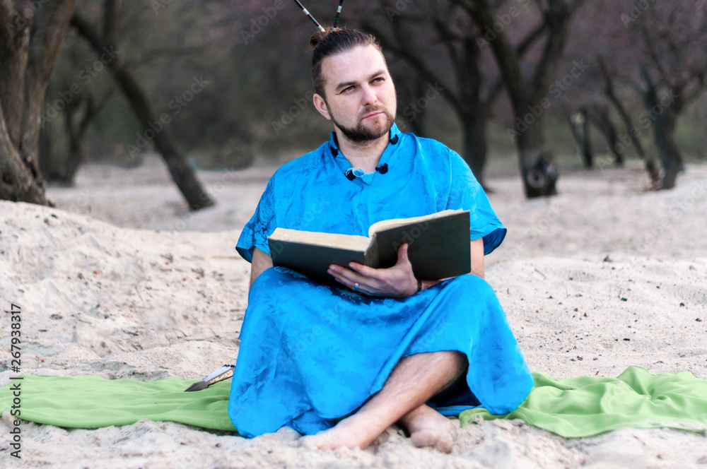 Obraz premium serious handsome bearded man in blue kimono sitting, holding opened large book and looking away on sandy beach near trees at spring