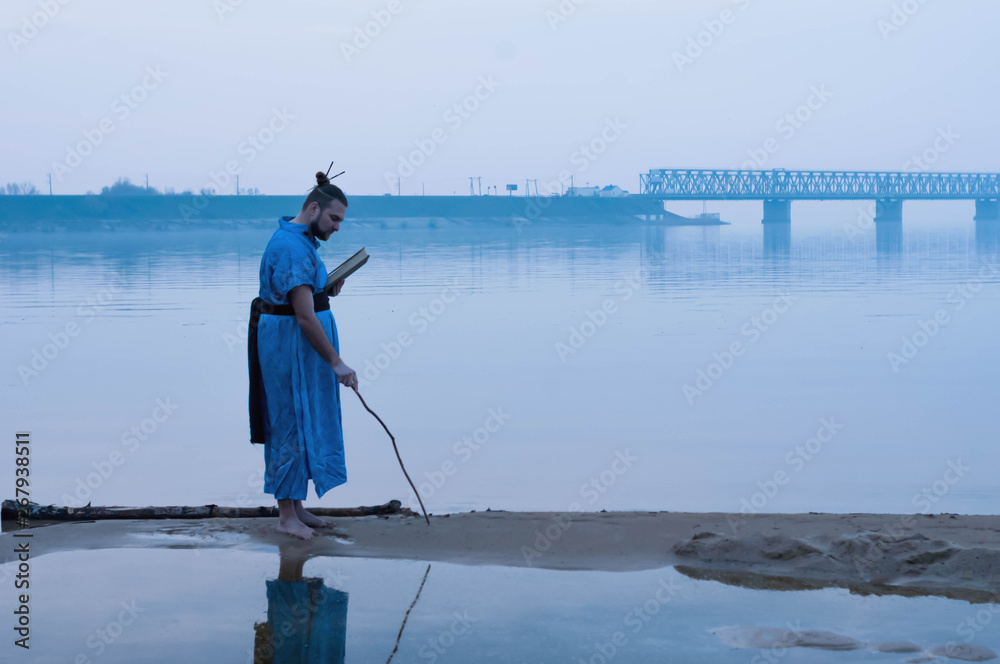 Naklejka premium side view of overweight bearded man in blue kimono standing on river bank and writing on sand with wooden stick and holding book in front of bridge
