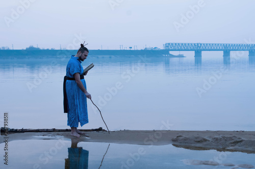 side view of overweight bearded man in blue kimono standing on river bank and writing on sand with wooden stick and holding book in front of bridge