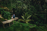 Couple standing on wooden bridge near rice terraces in Bali Indonesia. Holding hands. Romantic mood. Tropical vacation. Aerial shot. On background coconut palm trees.