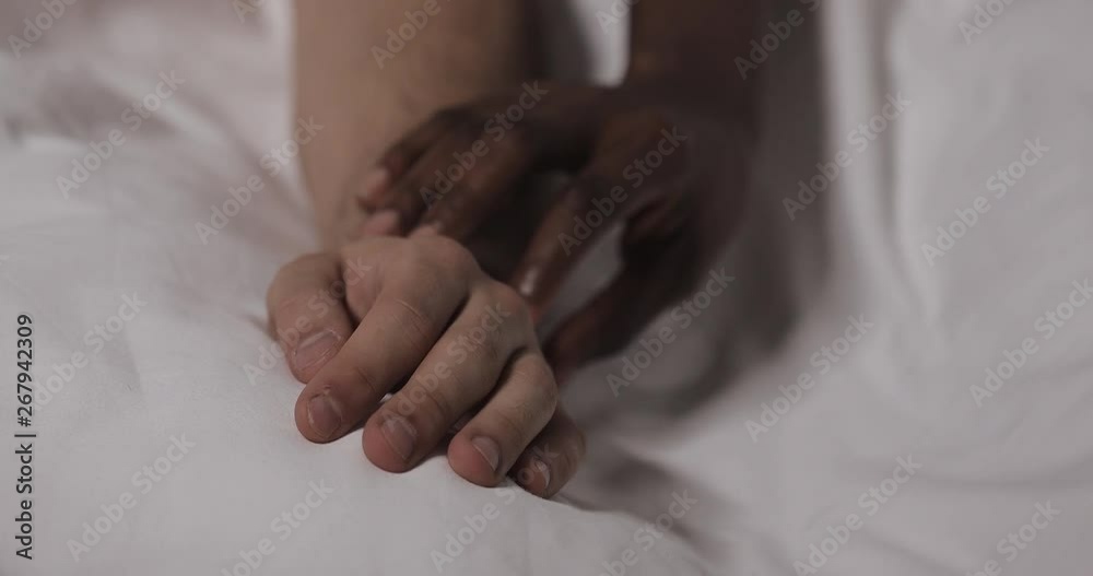 Multi-ethnic couple in love lying in bed together. Close up. Mans hand is lying on a bed, womans hand is caressing it and grabbing. Multi-ethnic couple in love lying in bed together. Close up. Mans hand is lying on a bed, womans hand is caressing it and grabbing.