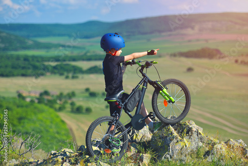 Little child stand next to his mountain bike on mountains edge and looks at the beautiful scenery