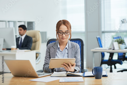 Young businesswoman in eyeglasses sitting at her workplace and concentrating on her work she using digital tablet for online work with businessman working in the background
