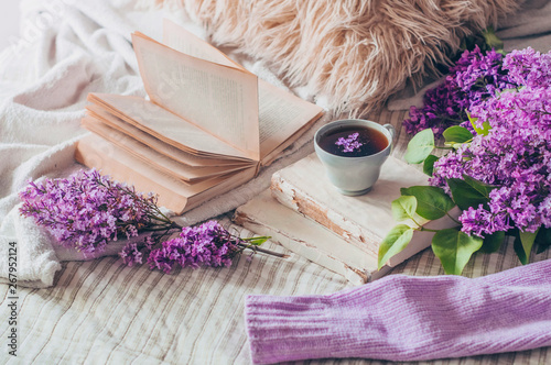 Fototapeta Naklejka Na Ścianę i Meble -  Still life details in home interior of living room. Sweaters and cup of tea with lilac flowers and spring decor on the books. Read, Rest. Cozy spring concept.