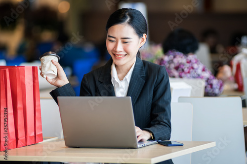 Young beautiful female wears black business suit works, shops online via cell phone and laptop with happy and smile face in department store. Business person, modern lifestyle, and shopping concept.