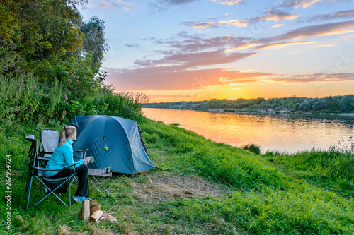 Young woman enjoys the sunset in the travel chair next to the tent in camping near the river