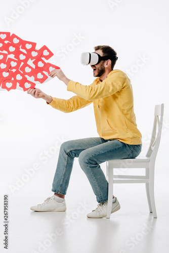 excited man in virtual reality headset sitting on chair and holding red paper cut cards with herts symbols on white background photo