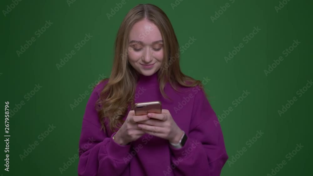 Beautiful female ginger student working with phone being attentive smiles into camera on green chroma background. Beautiful female ginger student working with phone being attentive smiles into camera on green chroma background.