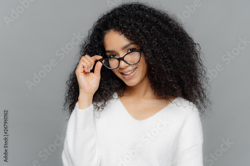 Horizontal shot of satisfied female teacher keeps hand on frame of glasses, smiles happily, has Afro hairstyle, wears casual white jumper, stands indoor over grey background. Good mood and beauty