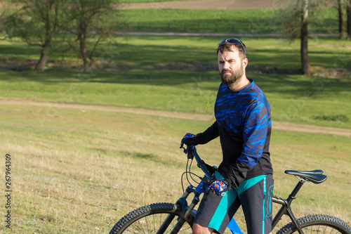 Cyclist in shorts and jersey on a modern carbon hardtail bike with an air suspension fork rides off-road on green hills near the forest 