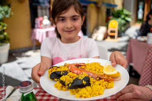 A young girl is offering a dish of typical spanish paella. Outdoor shot, the girl is blurred