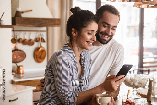 Photo of smiling brunette couple drinking coffee and using cell phone during breakfast at home
