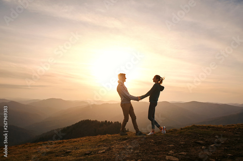 Silhouette of loving couple standing on edge of mountain and holding hands on sunset sky and mountains background.