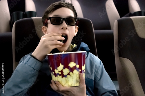 The teenager in the cinema in 3D glasses with popcorn in their hands. The guy opened his mouth and watches the movie in surprise.