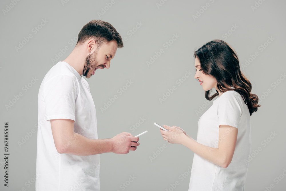 Fototapeta premium handsome man using smartphone and yelling while standing near displeased girl with smartphone isolated on grey