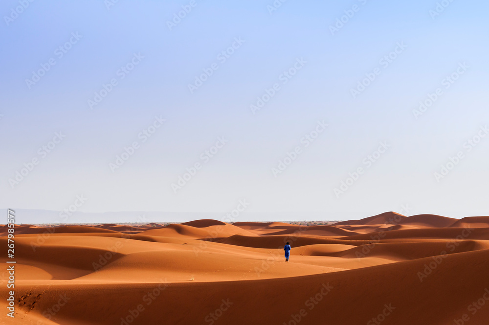 A person walks through sand dunes in the Sahara / A person in traditional clothing walks through the sand dunes in the Sahara, Morocco, Africa.
