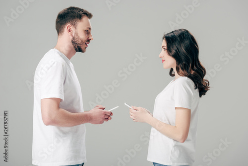 young man and woman in white t-shirts looking at each other while holding smartphones isolated on grey