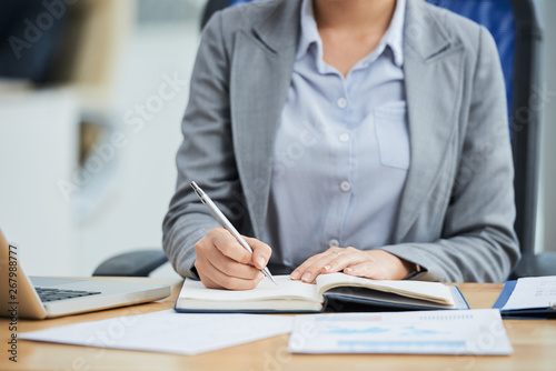 Close-up of young businesswoman sitting at the table planning her work day and writing it in notepad