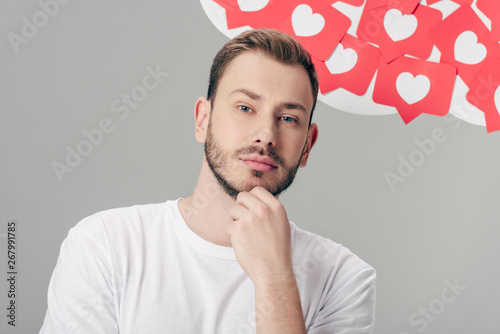 pensive handsome man in white t-shirt looking at camera near red paper cut cards with hearts symbols isolated on grey