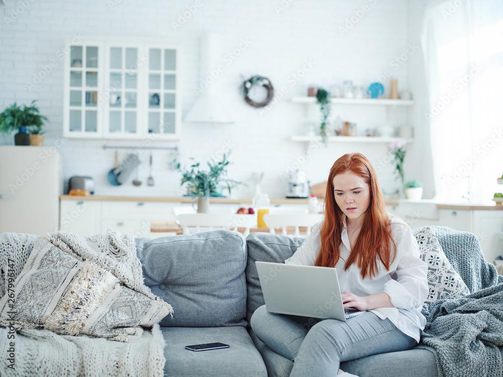 Beautiful young red-haired woman sitting on sofa at home and working on laptop looking confused Beautiful young red-haired woman sitting on sofa at home and working on laptop looking confused