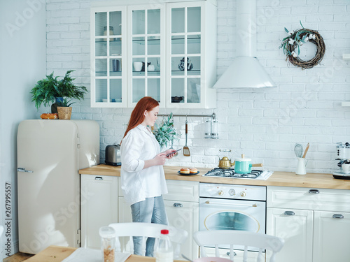Young woman text messaging on cell phone or browsing social networks while preparing breakfast in kitchen
