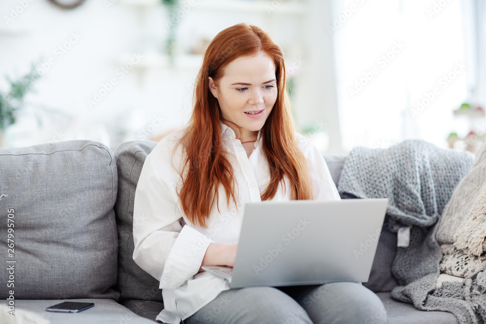 Young female blogger sitting on couch at home and working on laptop computer, her smartphone placed nearby Young female blogger sitting on couch at home and working on laptop computer, her smartphone placed nearby