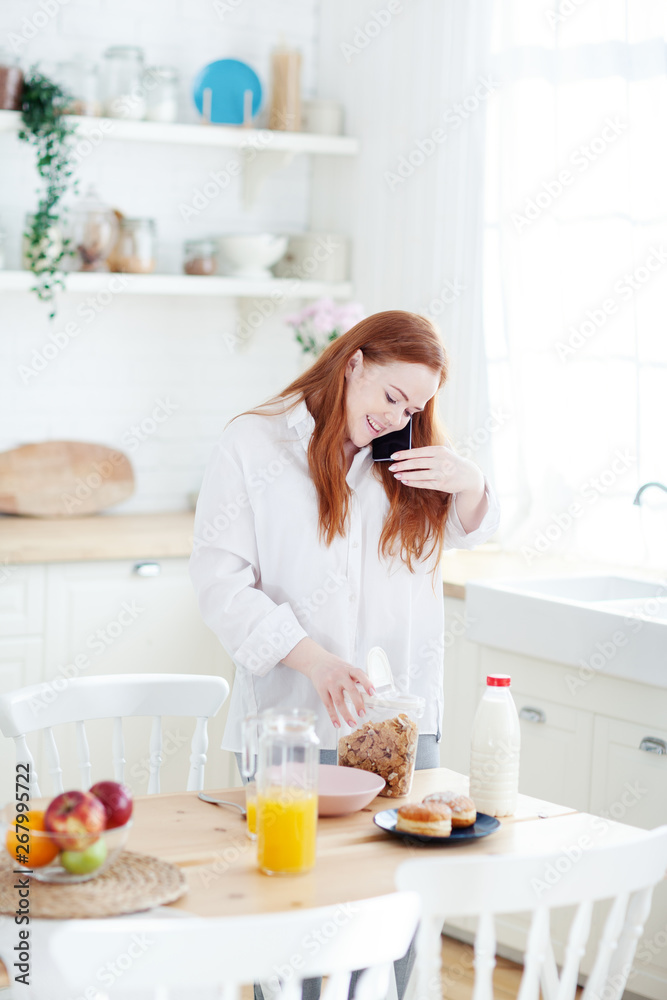 Smiling red-haired young woman talking on cell phone while preparing cereals with milk for breakfast in domestic kitchen.. Smiling red-haired young woman talking on cell phone while preparing cereals with milk for breakfast in domestic kitchen..
