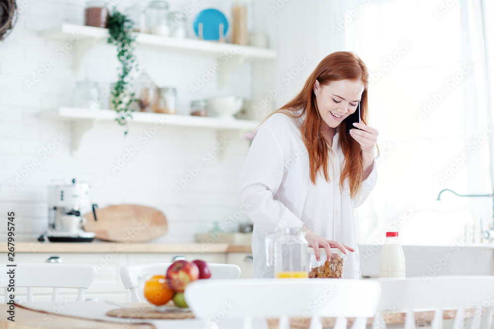 Cheerful young woman with red hair preparing breakfast of cereals, milk and orange juice and talking on cell phone standing at table in kitchen