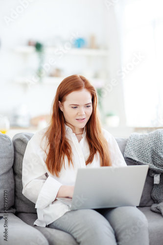 Authentic beautiful businesswoman with red-hair sitting on sofa at home and typing on laptop computer