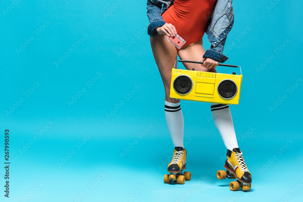 cropped view of woman in swimsuit and roller skates holding boombox and cassette tape on blue