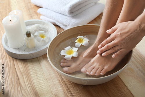 Closeup view of woman soaking her feet in dish with water and flowers on wooden floor. Spa treatment