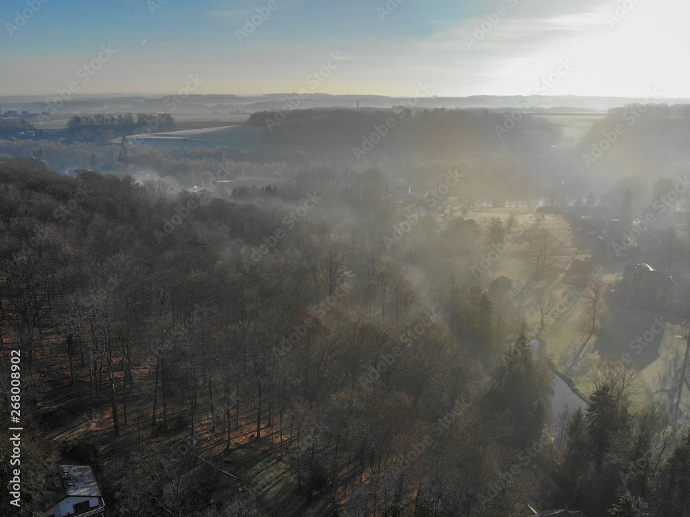 Aerial view of foggy and cold winter morning with blue sky facing the sun over the forest and farmland in Belgium, Walloon Brabant