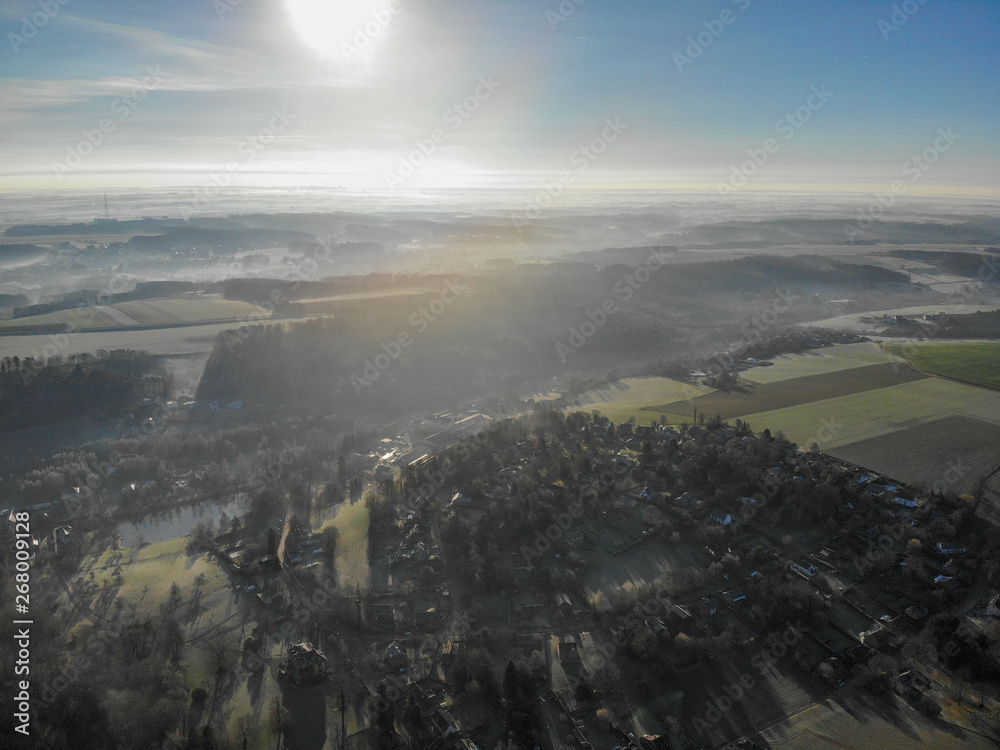 Aerial view of foggy and cold winter morning with blue sky facing the sun over the forest and farmland in Belgium, Walloon Brabant