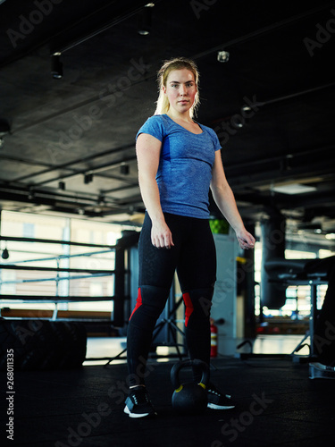 Full length three quarters portrait of determined woman wearing knee supporters standing near kettlebell and looking at camera before exercise in gym