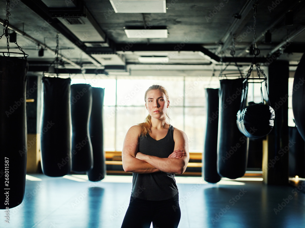 Three quarter length portrait of powerful athletic woman standing with her arms crossed among boxing bags in gym and looking at camera