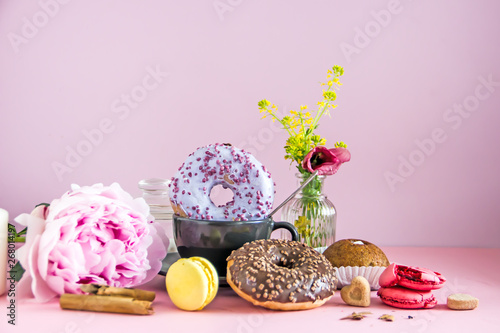 A black cup of coffee, donuts in colored glaze, a bunch of flowers, macaroons, cinnamon sticks, sugar bowl, cakes stand on a pink background. The concept of a sweet breakfast, save space.
