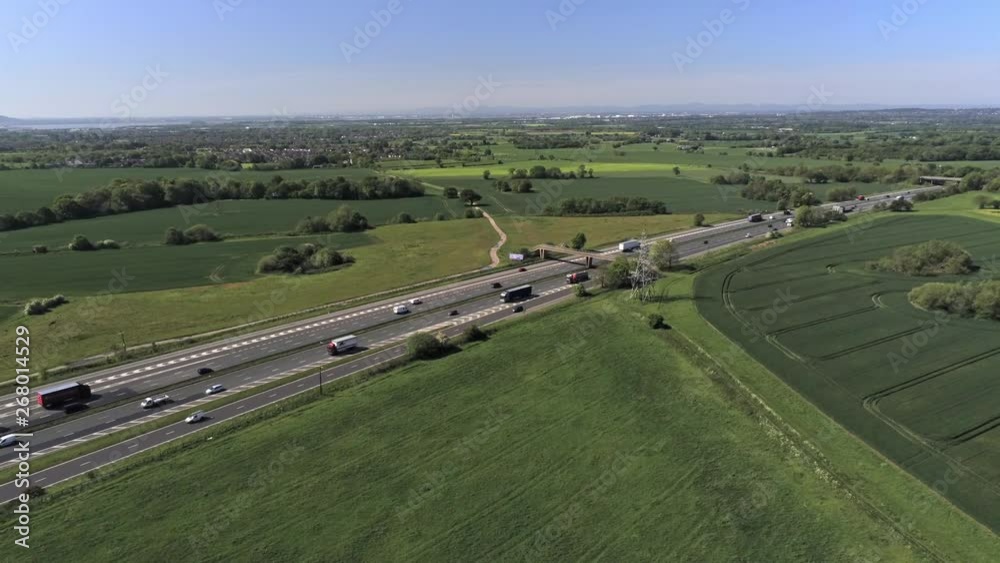 Static aerial view overlooking M62 motorway with rush hour morning traffic surrounded by sunny British farmland countryside off into the distant skyline.
