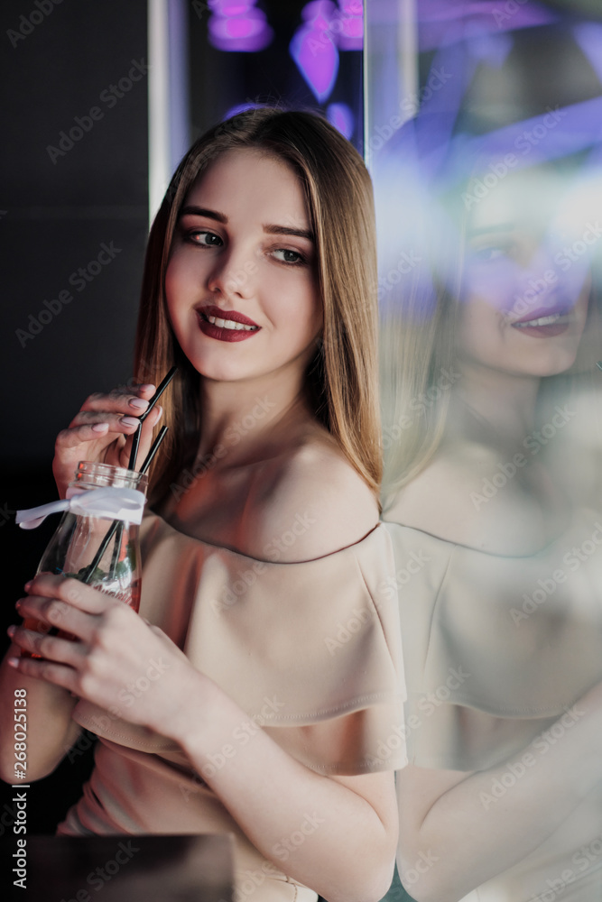 Young beautiful brown-haired girl in a nightclub with neon lights sits by the glass and drinks a red cocktail. Young beautiful brown-haired girl in a nightclub with neon lights sits by the glass and drinks a red cocktail.