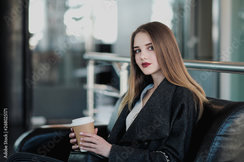 A young beautiful brunette girl in a gray coat is sitting on a bench waiting. Drinking coffee and holding a paper cup with a drink.