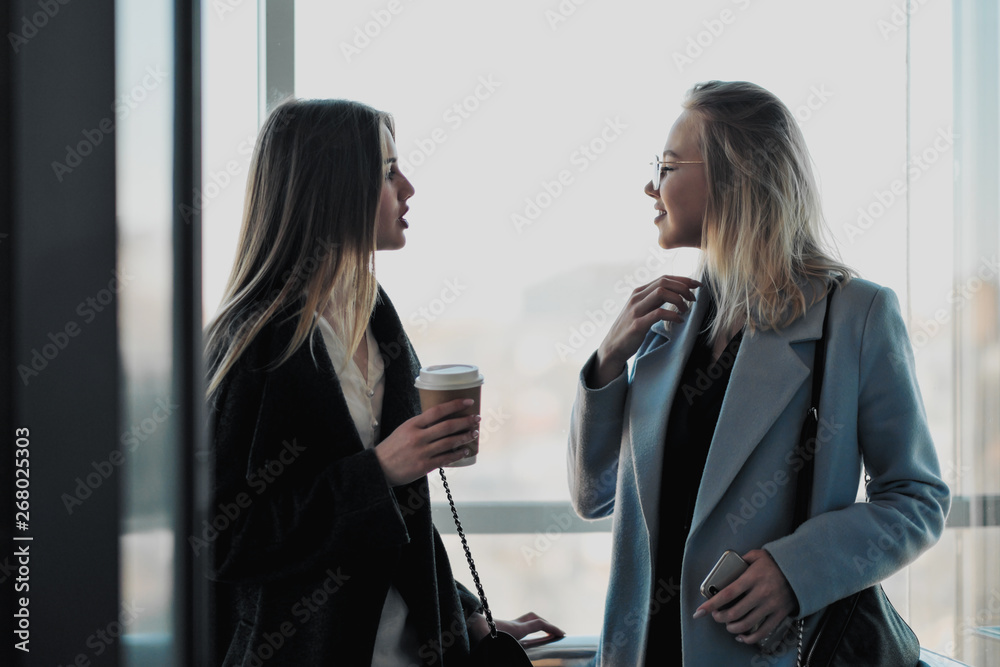 Two beautiful girls, a blonde and a brunette, are wearing a coat in a glass elevator with a window. Shopping center or office in the city. Two beautiful girls, a blonde and a brunette, are wearing a coat in a glass elevator with a window. Shopping center or office in the city.