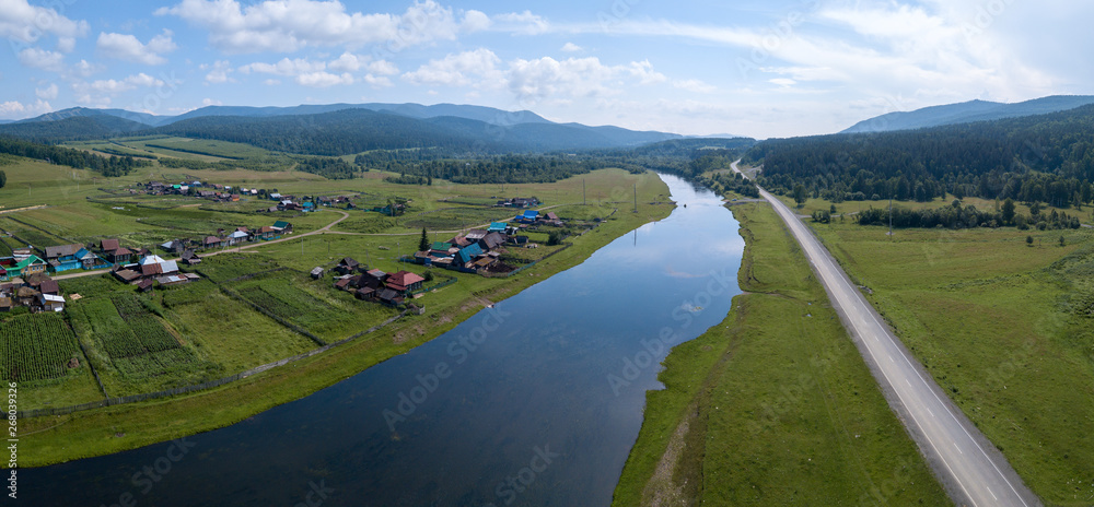 Aerial panorama of the river of Belaya with the village on its green coast, asphalt road along the river and Ural Mountains on the horizon