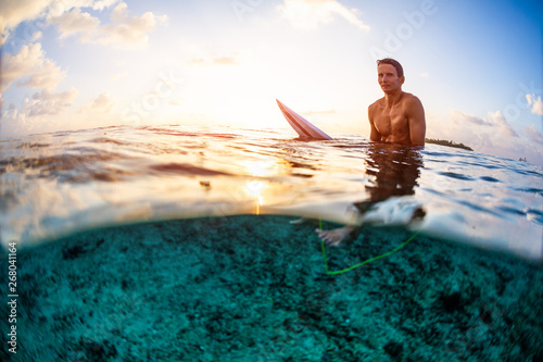 Surfer sits on the surfboard and looks at the camera at sunset. Splitted view with underwater view © Dudarev Mikhail